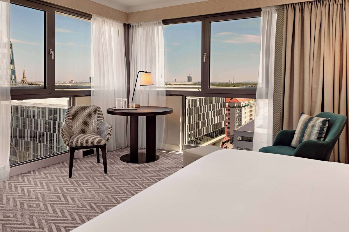 A hotel room with a large bed, two chairs, a small table, and a view of the cityscape.