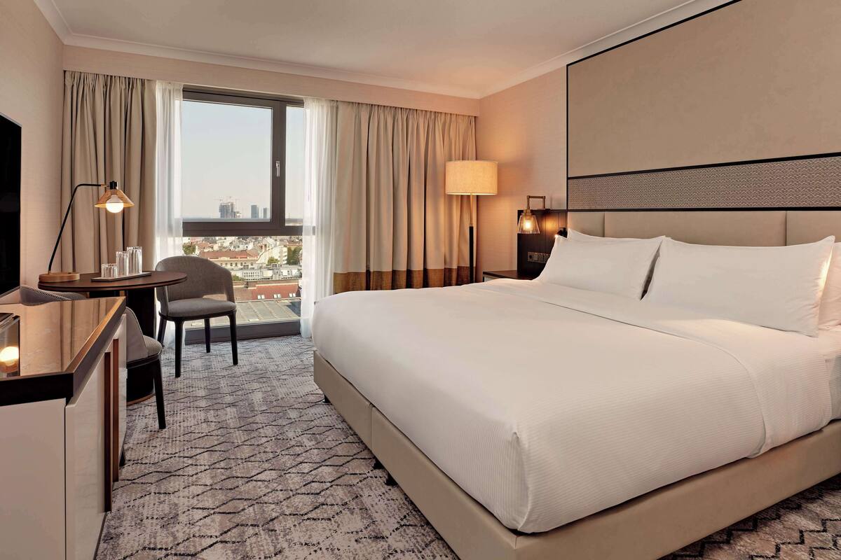 A hotel room with a large bed, a desk with a chair, a lamp, and a view of the city through the window.