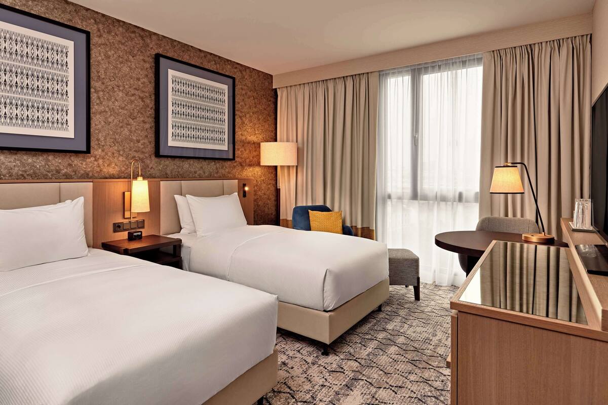 A hotel room with two beds, a desk, a chair, a TV, and a large window with curtains.