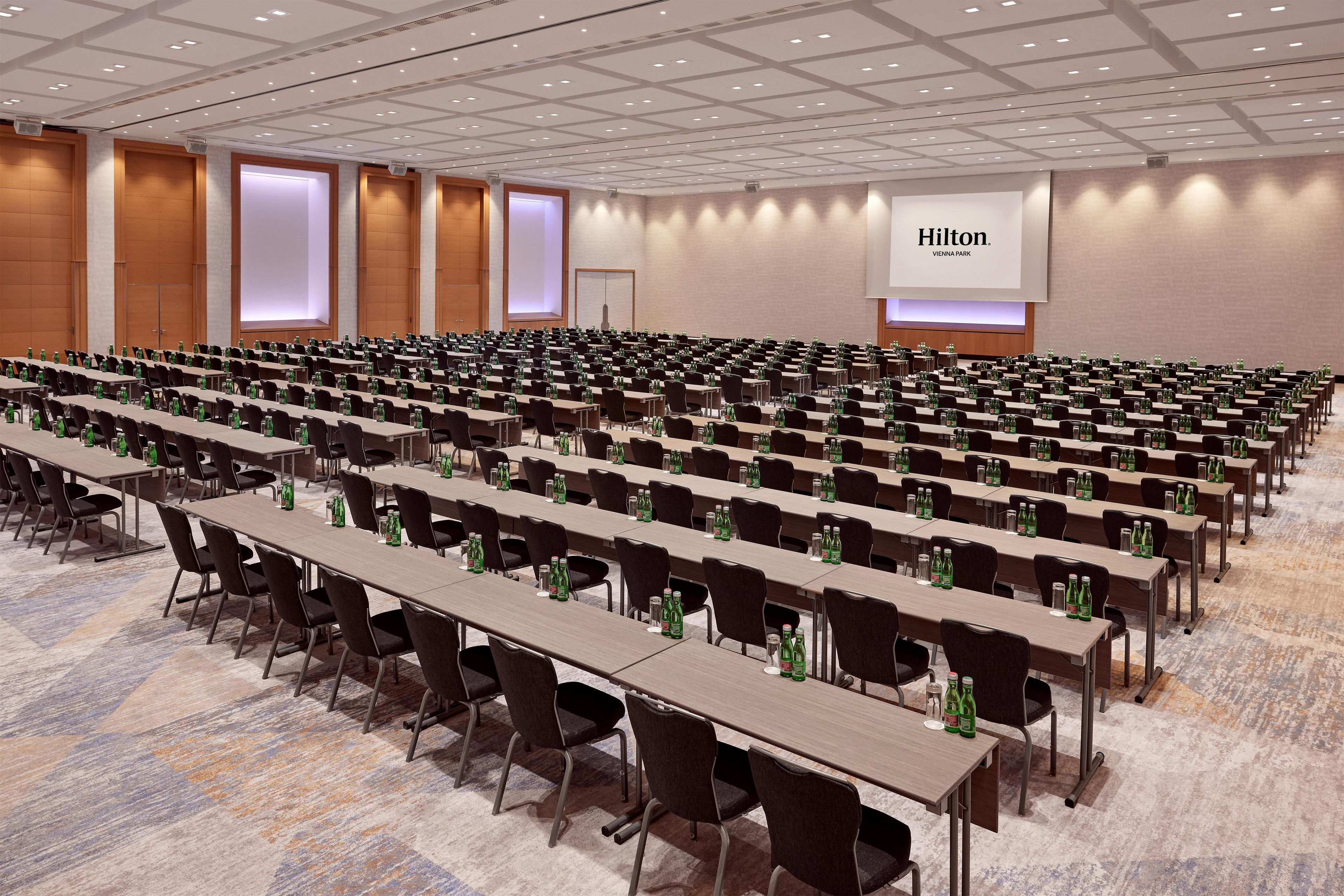 A large conference room with rows of tables and chairs, a large screen displaying 'Hilton Vienna Park', and a carpeted floor.