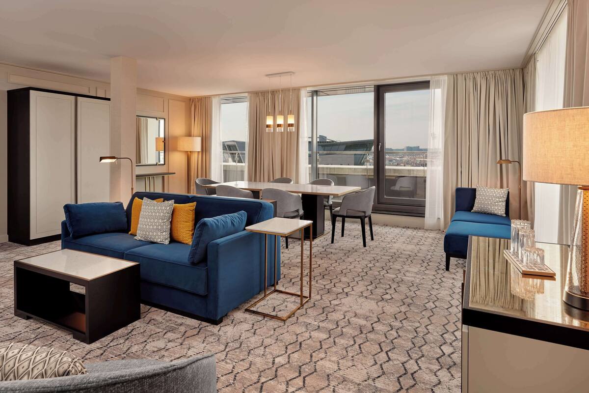 A modern hotel room with a blue sofa, a small table, a dining area, and a large window with a city view.