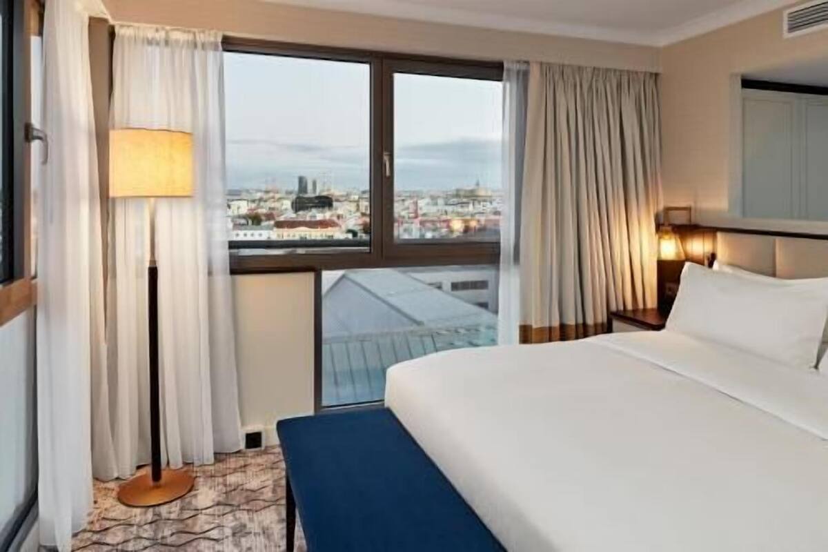 A hotel room with a bed, a desk, a lamp, a view of a cityscape, and a window with curtains.