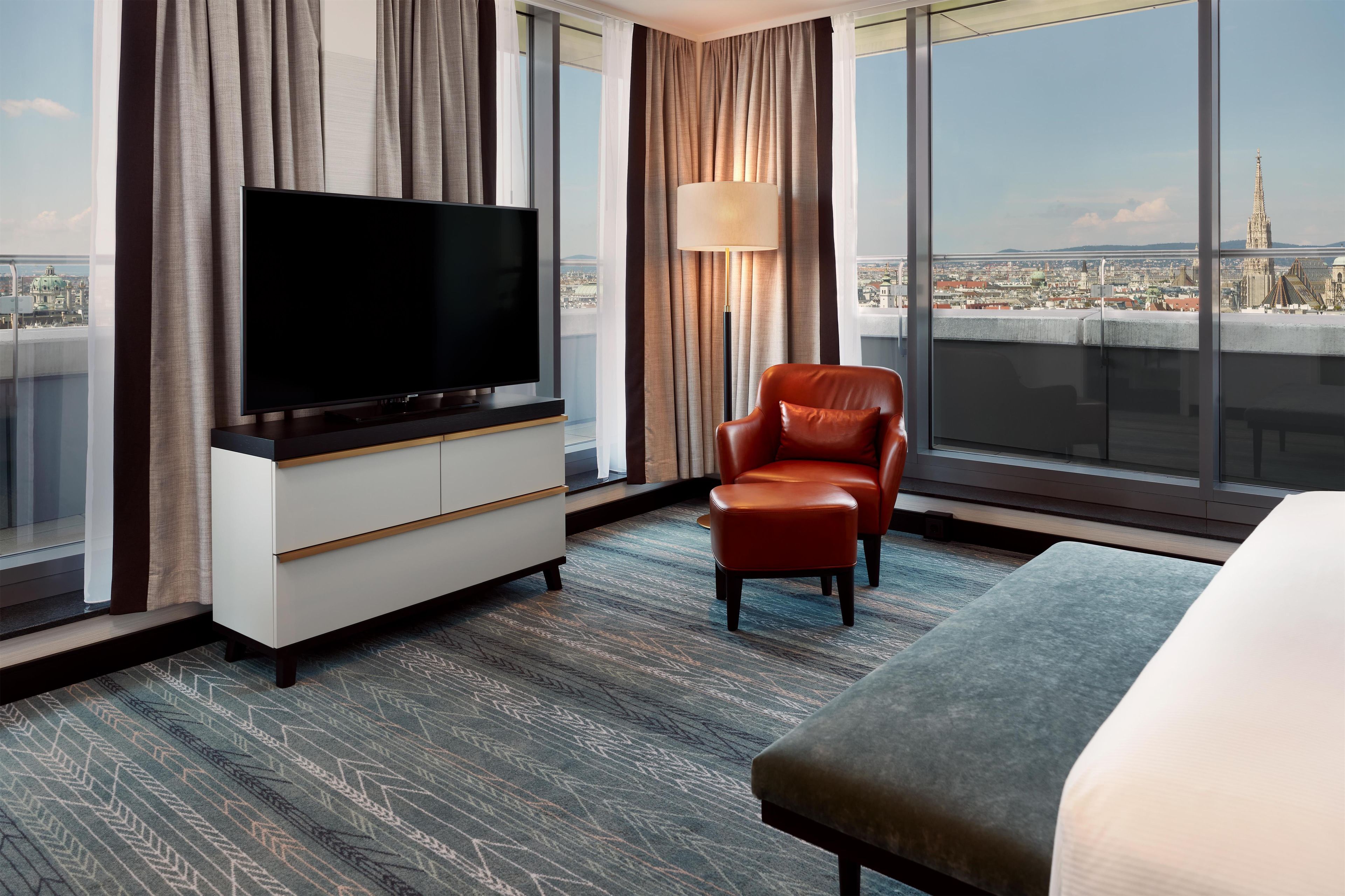 A hotel room with a large flat-screen TV, a leather armchair, a bed, and a view of a cityscape through large windows.