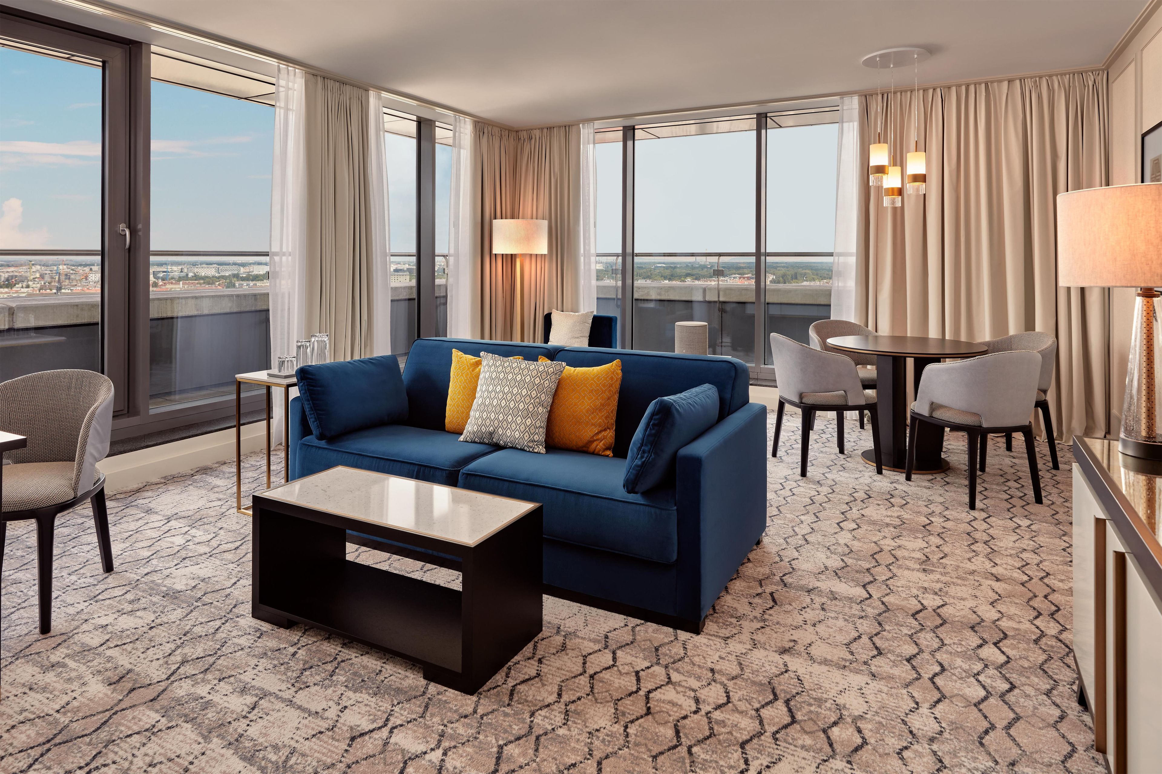 Habitación de hotel con un sofá azul, una mesa de centro, una mesa de comedor con sillas y ventanas amplias con vistas a la ciudad.