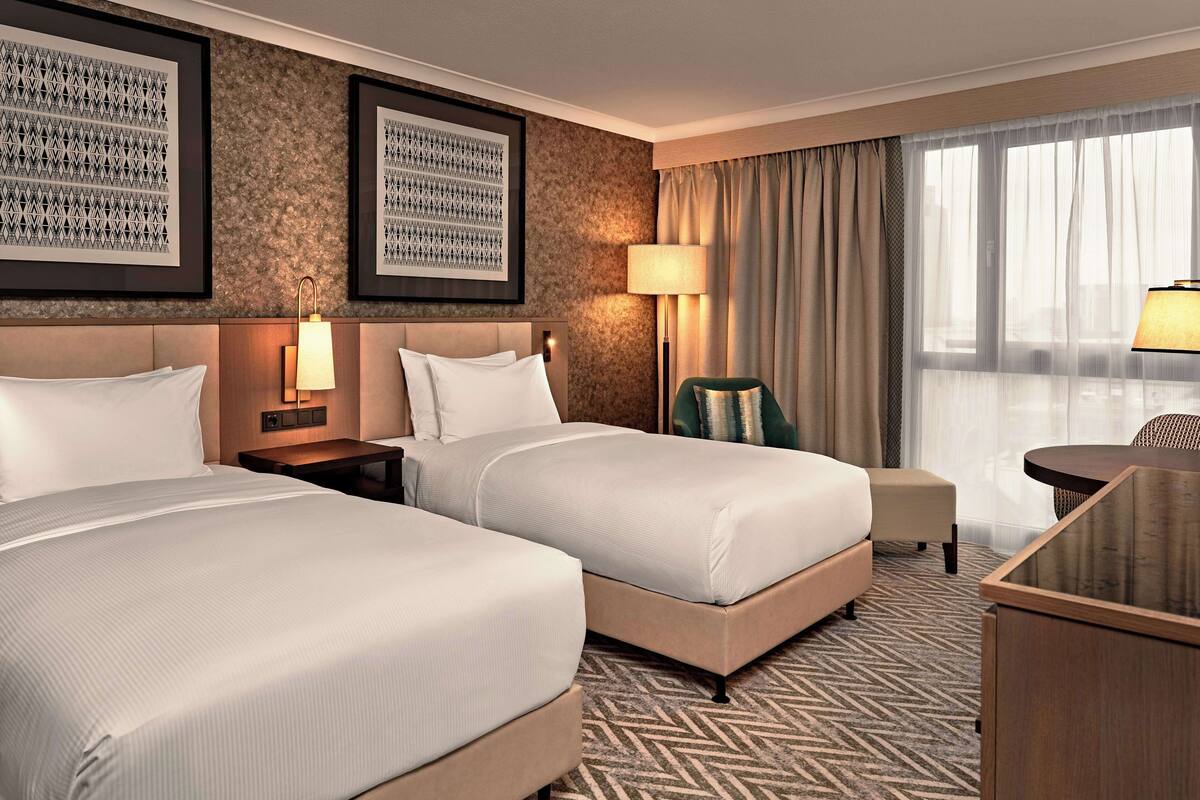 A hotel room with two beds, a desk, a chair, a lamp, and a large window with curtains.