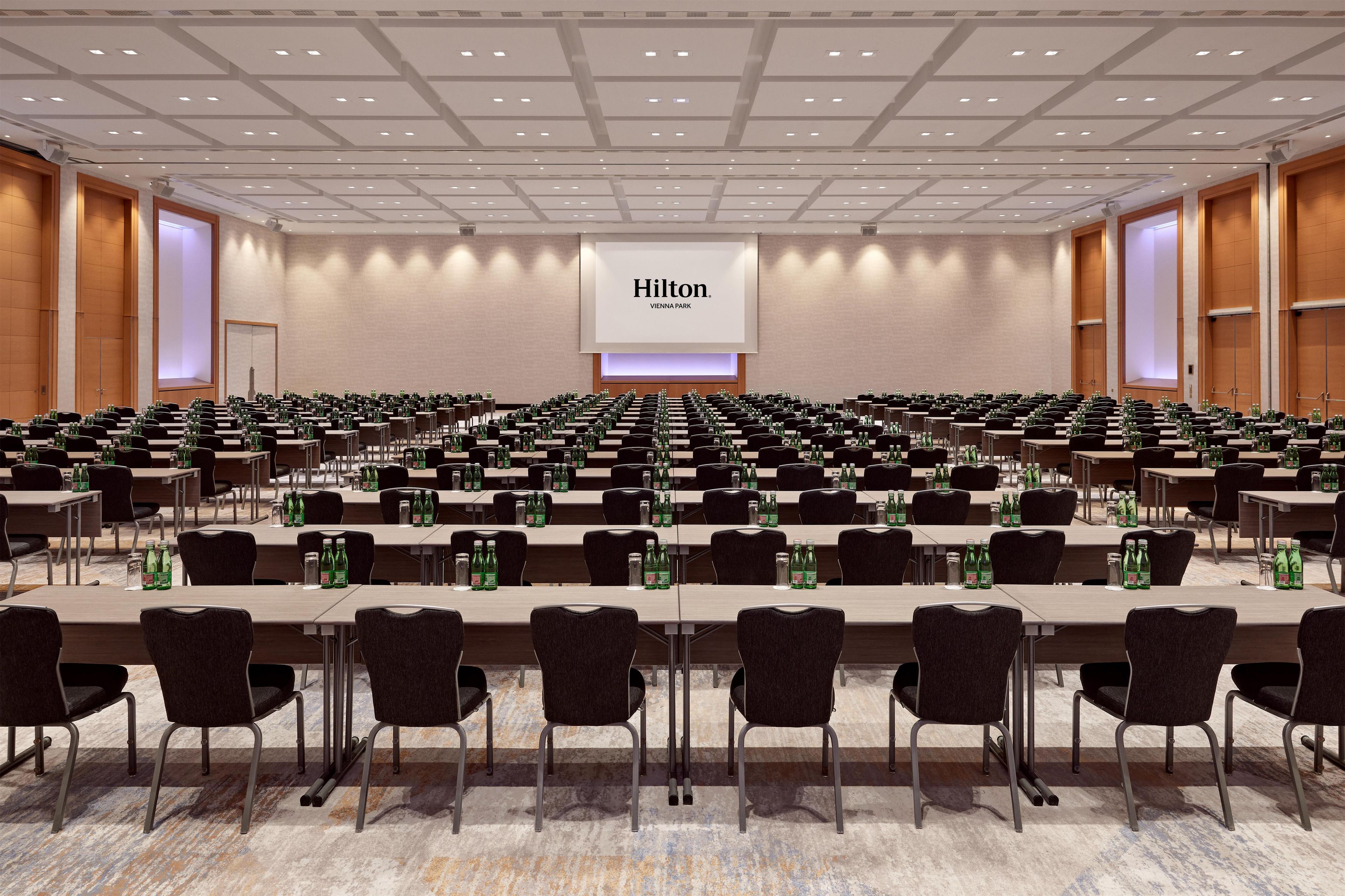 Un amplio salón de conferencias con filas de mesas y sillas, una gran pantalla mostrando 'Hilton Vienna Park', y una paleta de colores neutros.