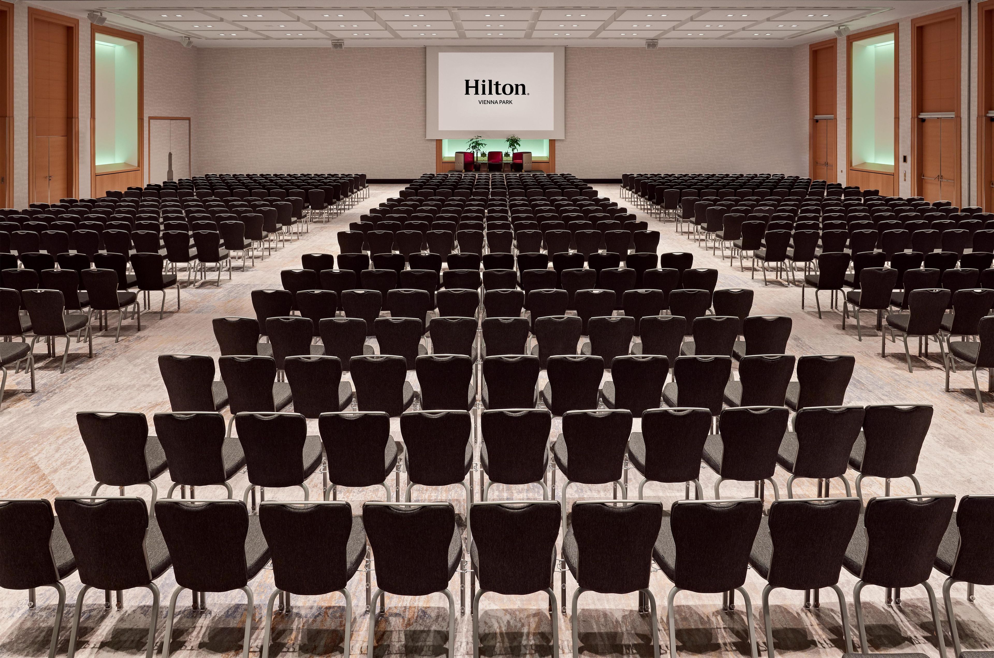 A large conference room with rows of black chairs, a podium at the front, and a large screen displaying "Hilton Vienna Park".
