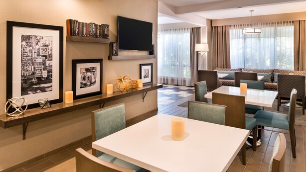 A hotel dining area with a white table, cushioned seating, a flat-screen TV, framed pictures, and a bookshelf. - Hampton Inn Los AngelesWest Covina (West Covina)