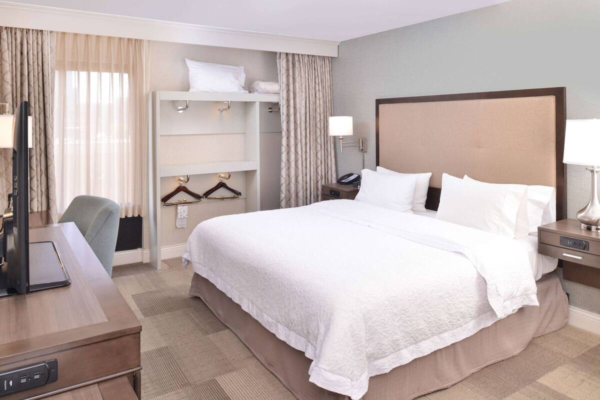 Een hotelkamer met een groot bed, een bureau met een computer, een stoel, een kledingkast en een nachtkastje met een lamp.