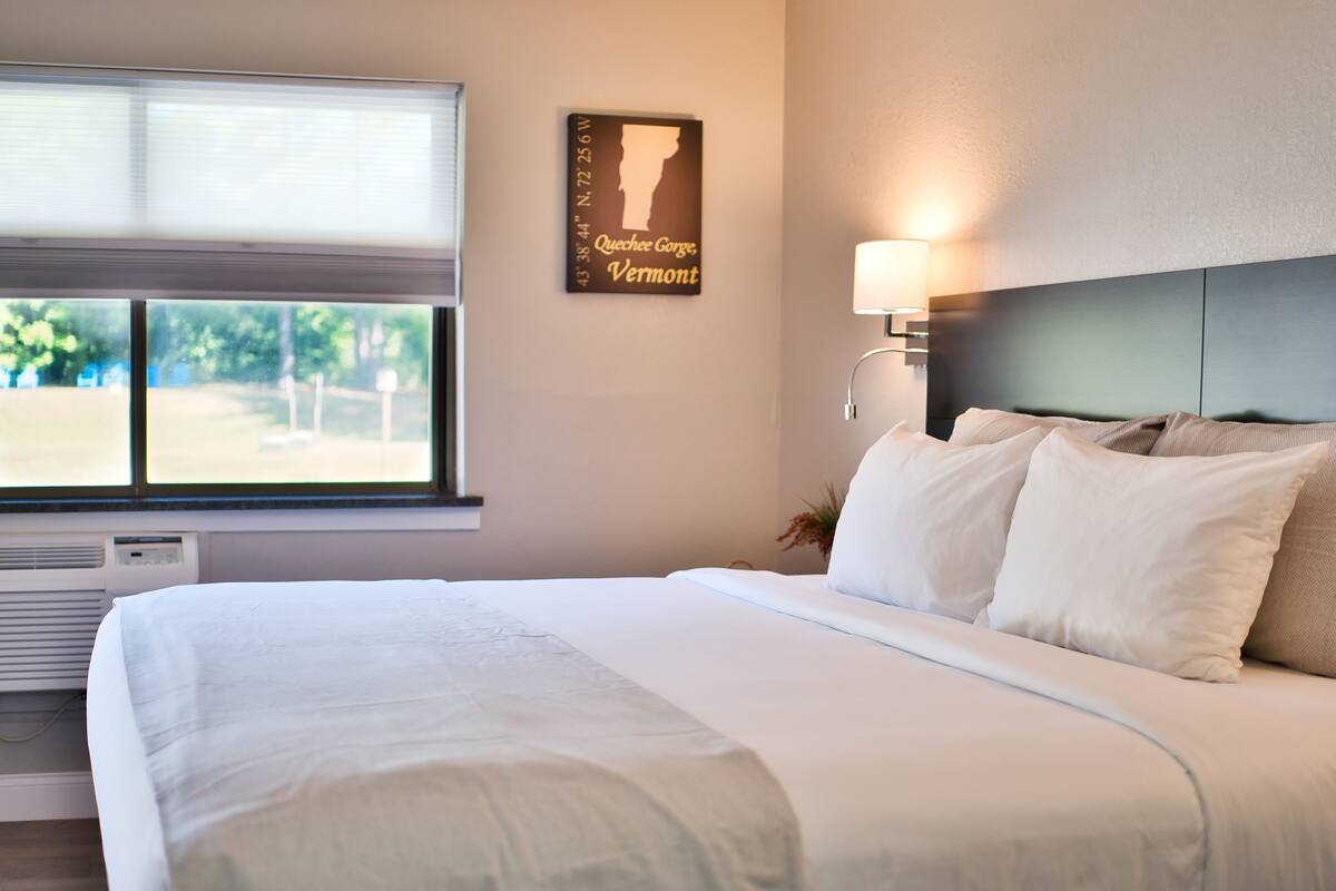 A neatly made bed with white linens, a wall-mounted map of Vermont, and a window with blinds.
