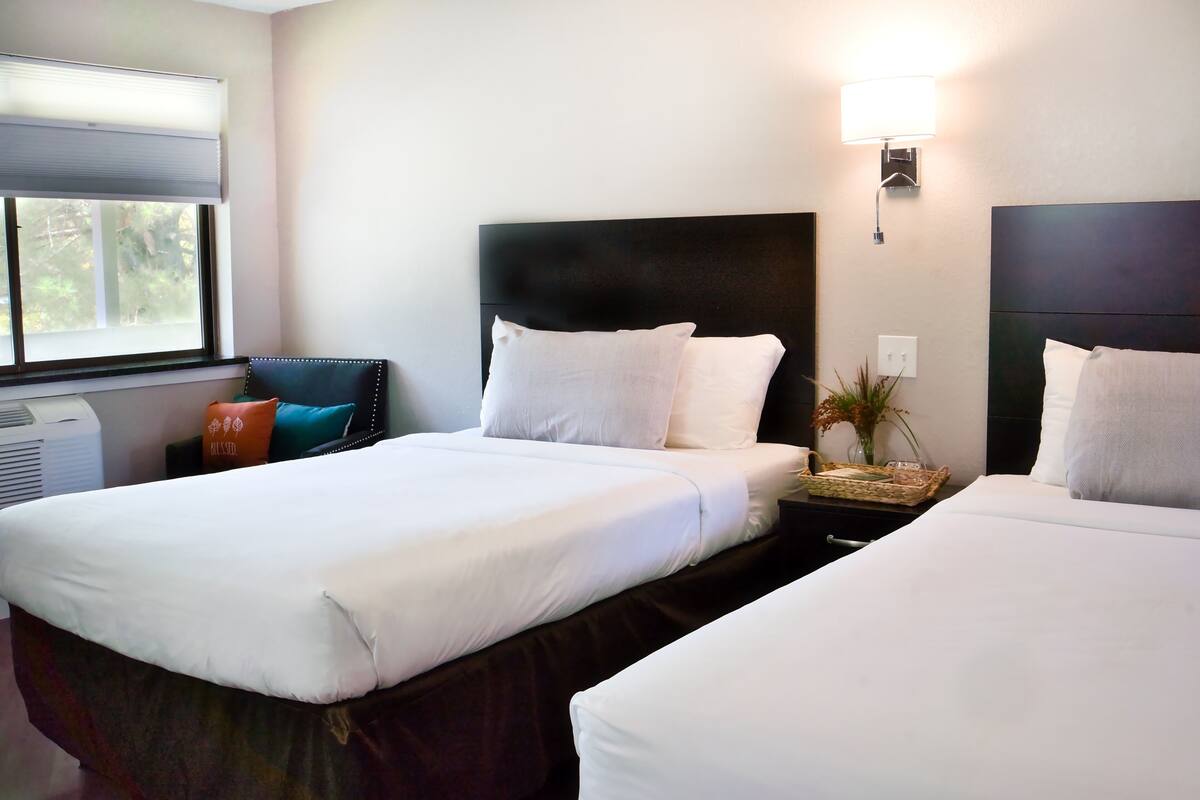 A hotel room with two beds, a nightstand, a lamp, and a window with blinds.