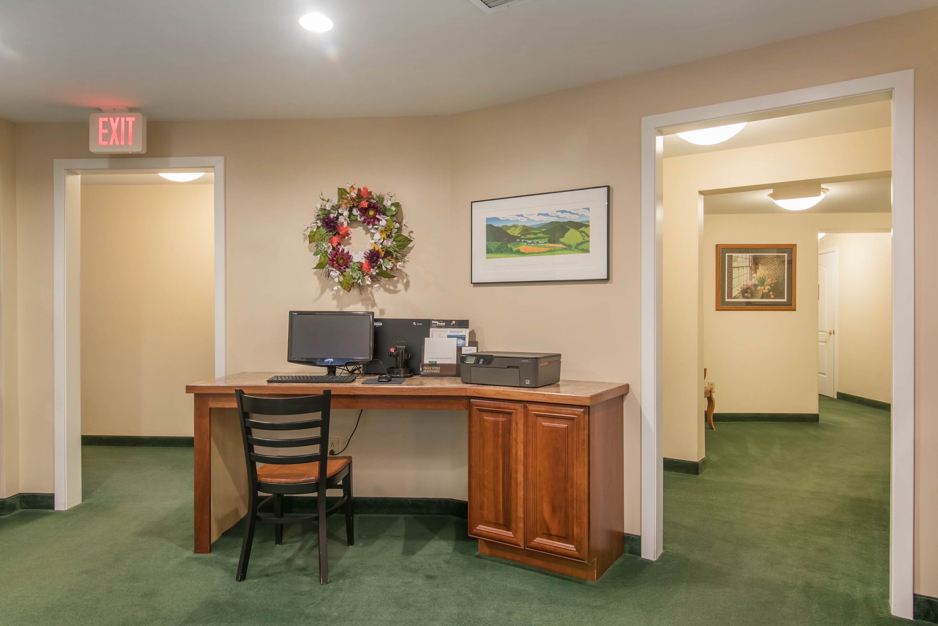 Habitación de hotel con un escritorio de madera, una computadora, una impresora, una silla, un cuadro enmarcado y una corona decorativa en la pared.