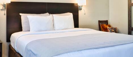 A neatly made bed with a dark headboard, two wall-mounted lamps, and a small wooden chair with a cushion.