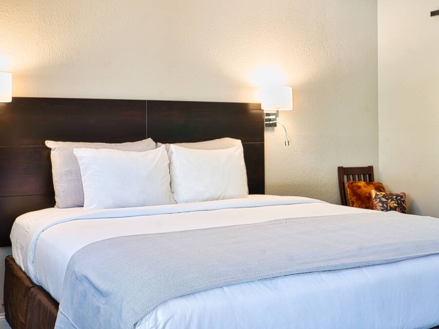 A neatly made bed with a dark headboard, two wall-mounted lamps, and a small wooden chair with a cushion.