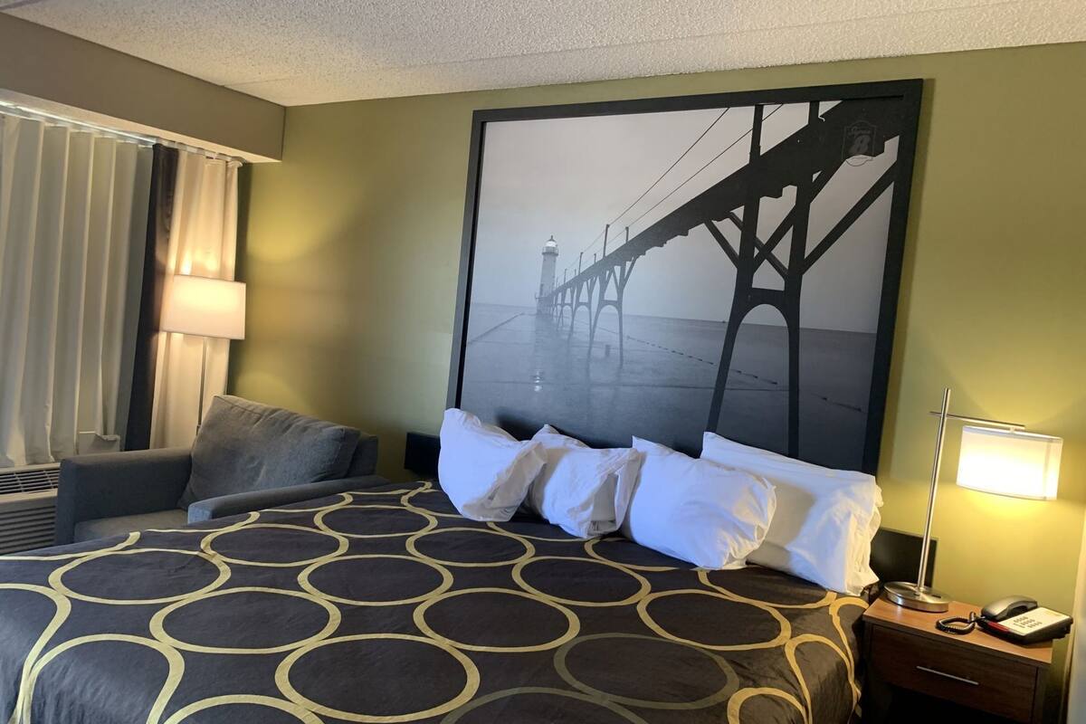 A hotel room with a large bed, a nightstand with a phone, a lamp, a chair, and a wall mural of a bridge.