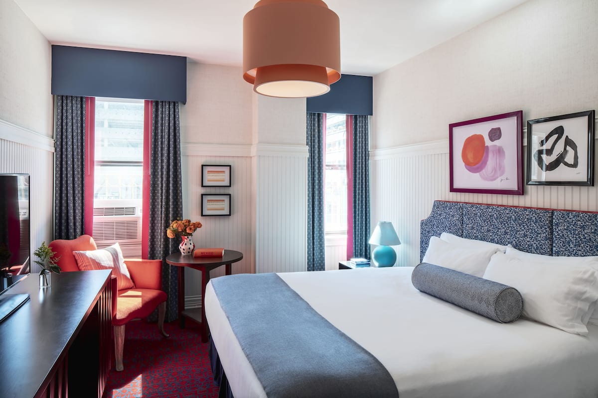 A hotel room with a bed, a desk, a chair, a lamp, and artwork on the wall.