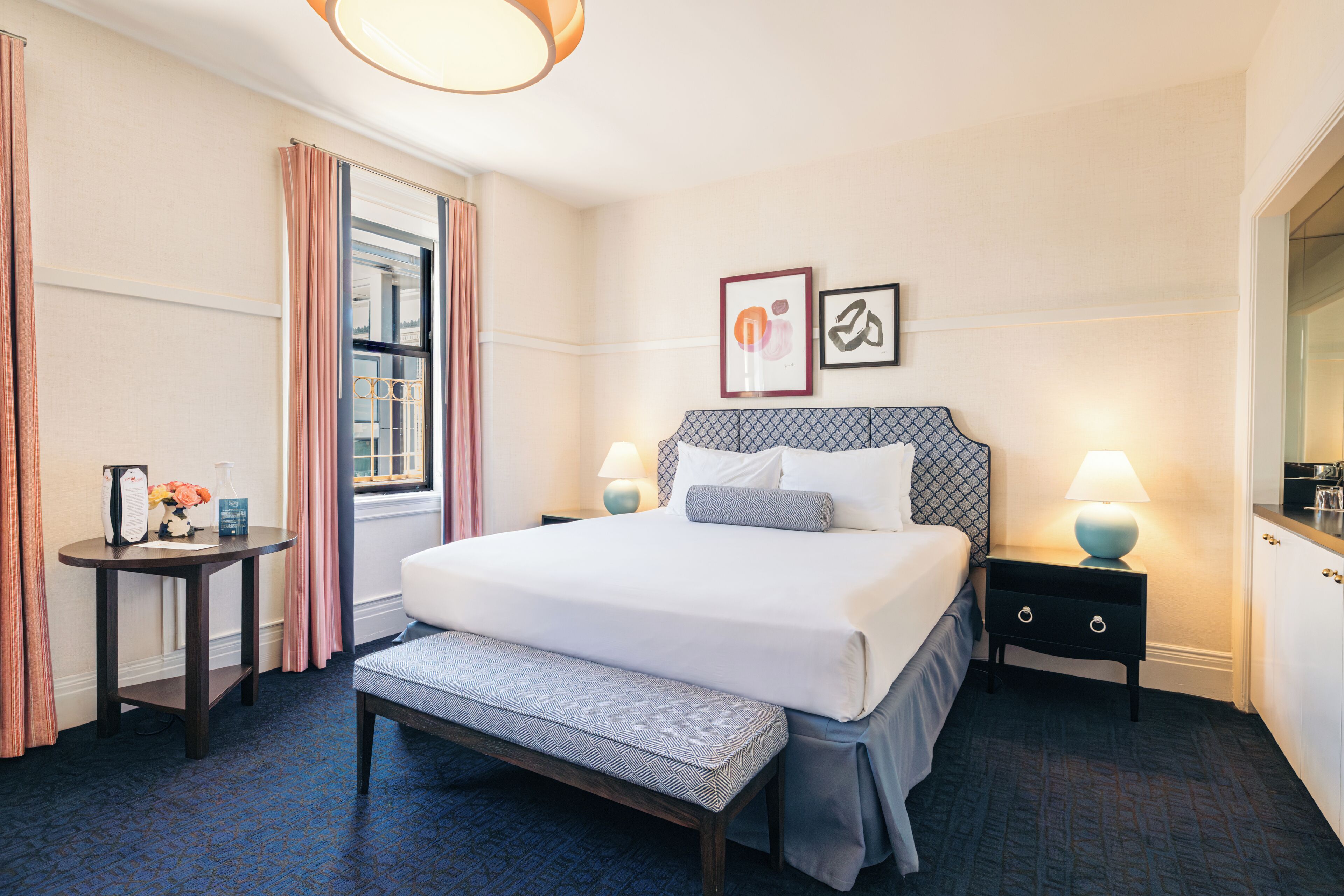 A hotel room with a large bed, bedside tables, a bench, a window with curtains, and artwork on the wall.