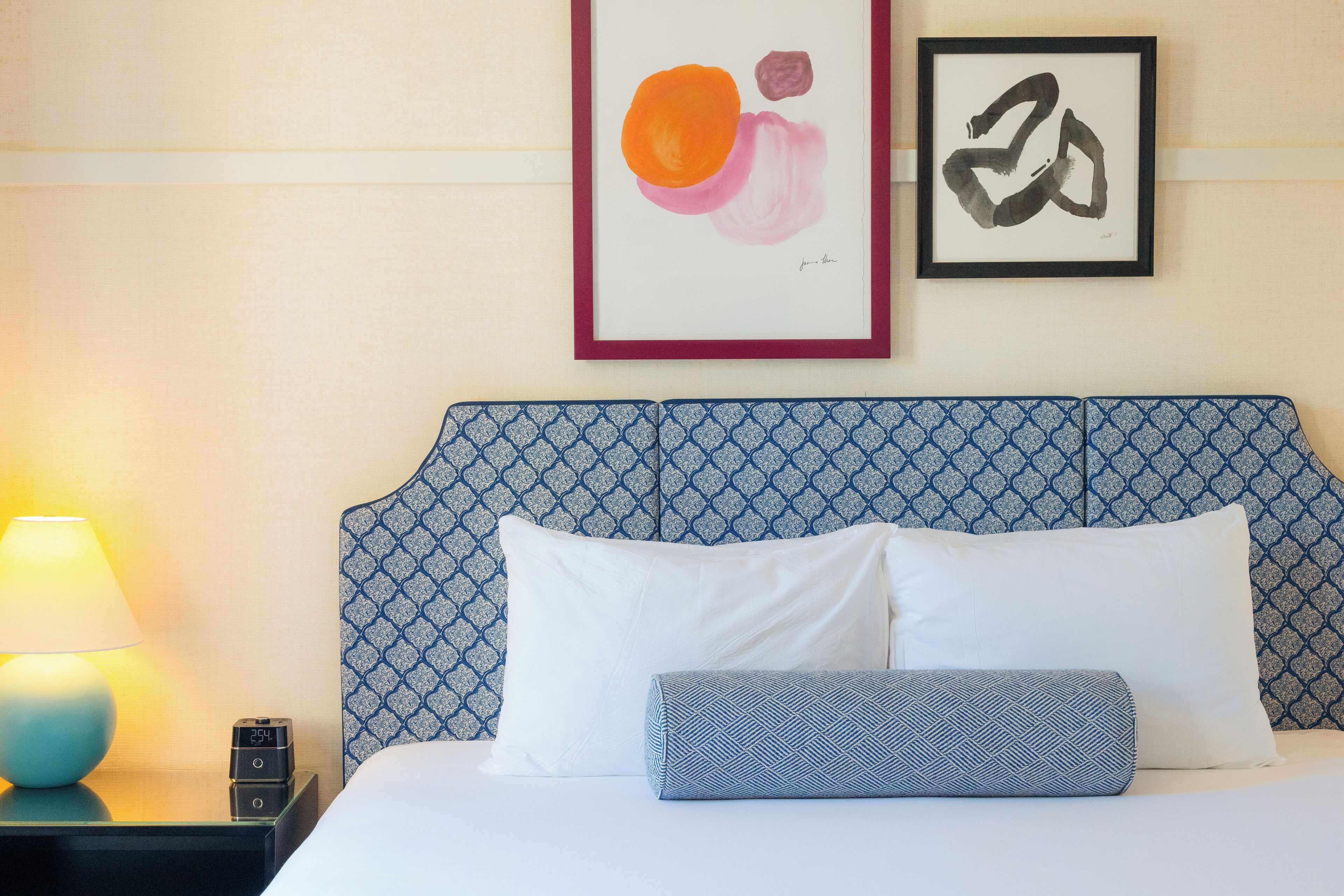 A neatly made bed with two pillows and a blue patterned pillow, a bedside lamp, and a framed abstract painting on the wall.