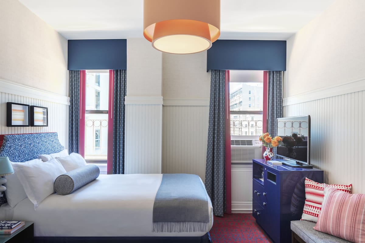 A hotel room with a bed, a desk, a chair, a dresser, and a window with curtains.