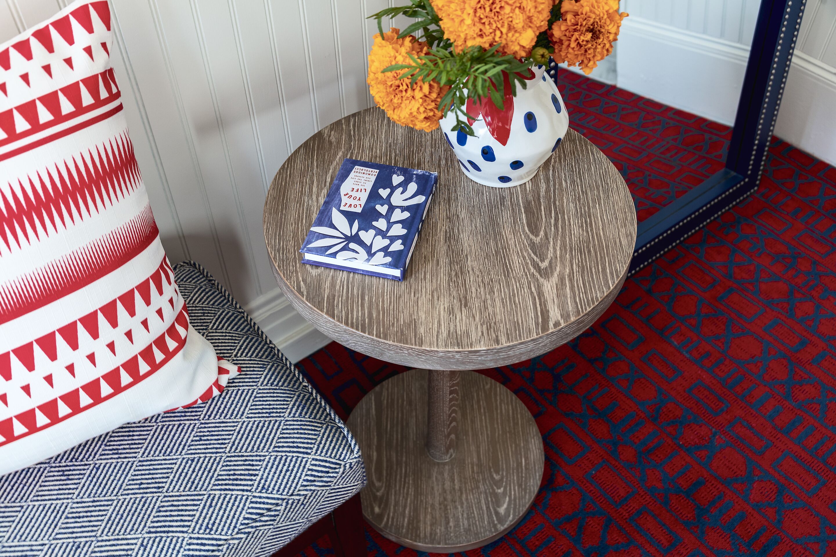 Une table ronde en bois, sur laquelle se trouvent un livre bleu et un vase blanc contenant des fleurs oranges.