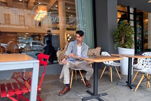 A man sitting at an outdoor cafe with a table and chairs, a potted plant, and a storefront in the background.