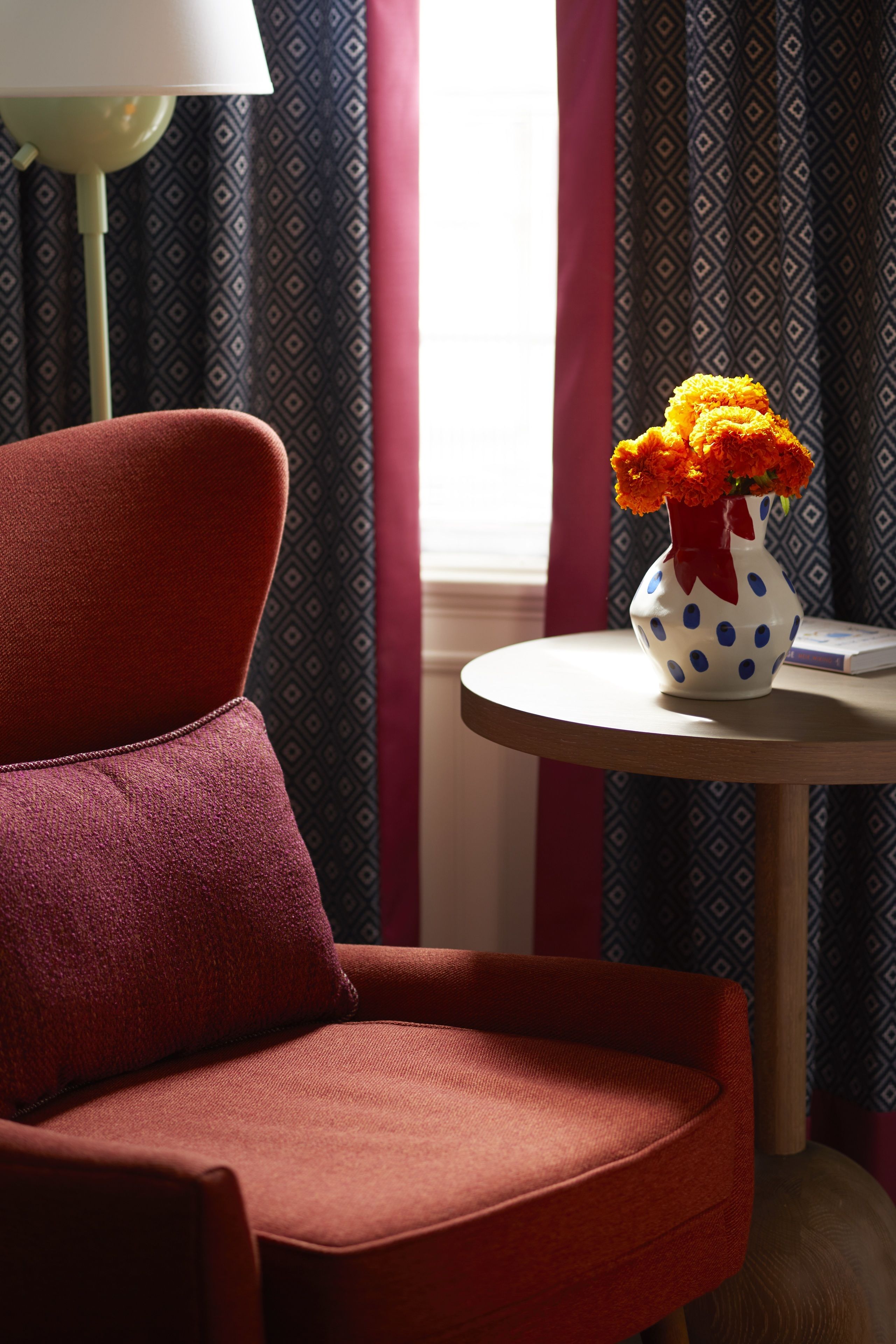 A red chair, a white vase with blue polka dots, and a patterned curtain in a room.