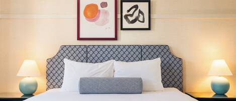 Una cama bien hecha con sábanas blancas, dos almohadas y una almohada estampada. Hay dos lámparas de pared a cada lado de la cama.