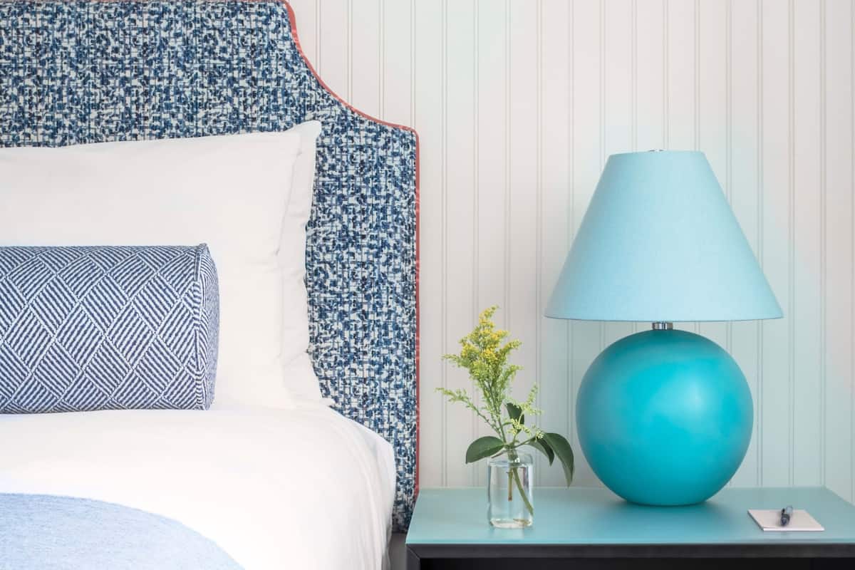 A neatly made bed with patterned bedding, a blue lamp on a bedside table, and a small vase with flowers.