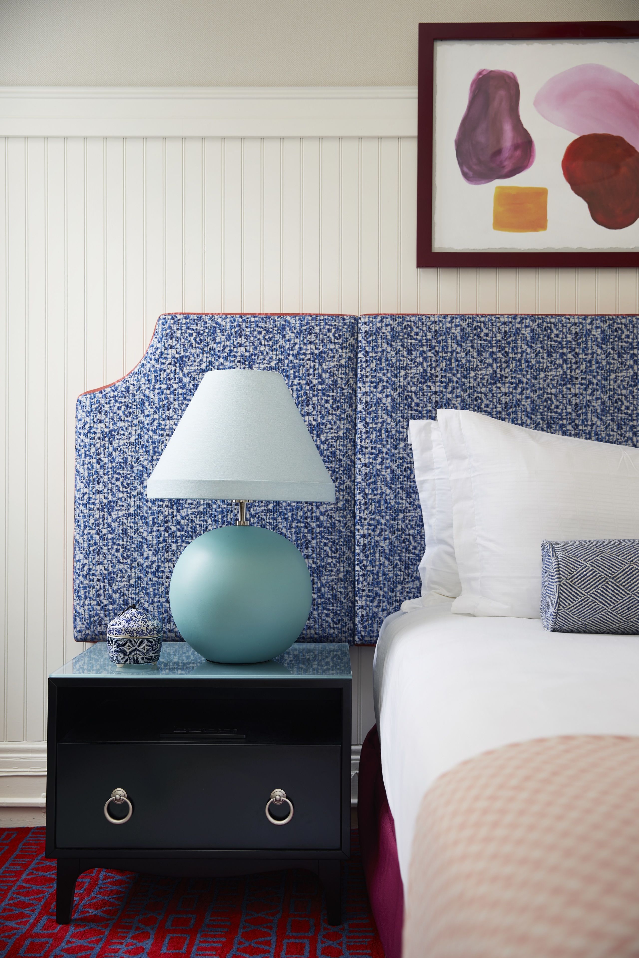 Un dormitorio ordenado con un cabecero azul con estampado, una cama blanca, una lámpara azul y un pequeño objeto decorativo en la mesita de noche.