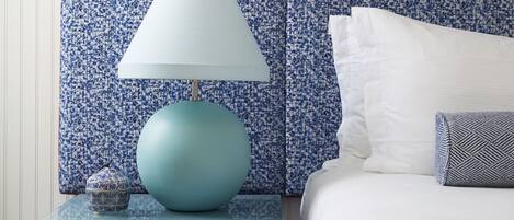 Un dormitorio ordenado con un cabecero azul con estampado, una cama blanca, una lámpara azul y un pequeño objeto decorativo en la mesita de noche.