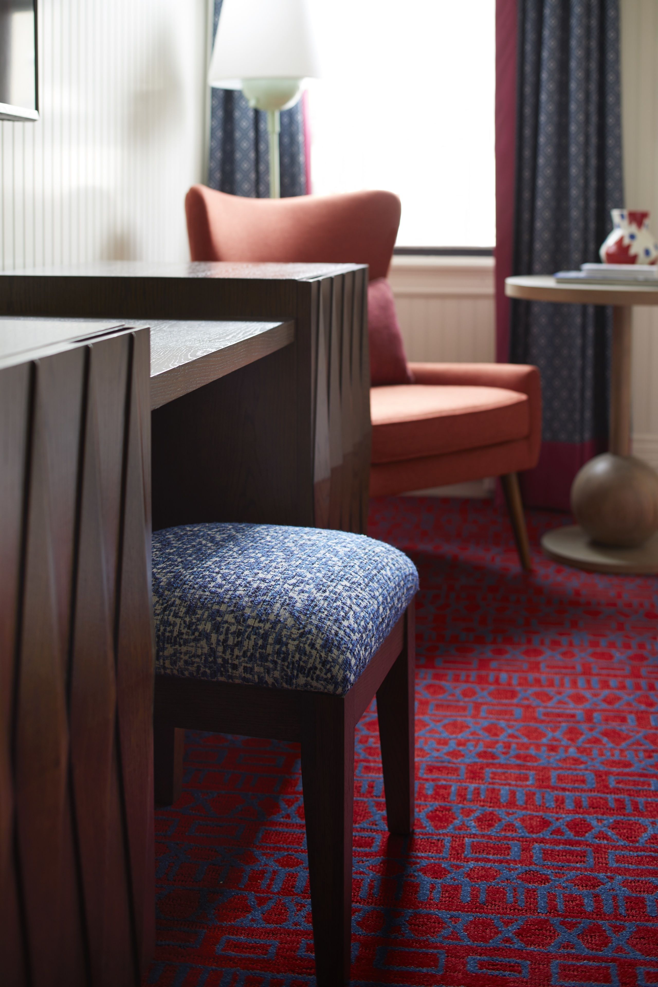 A hotel room with a wooden desk, a patterned blue chair, a red patterned carpet, a pink armchair, and a small round table with a globe.