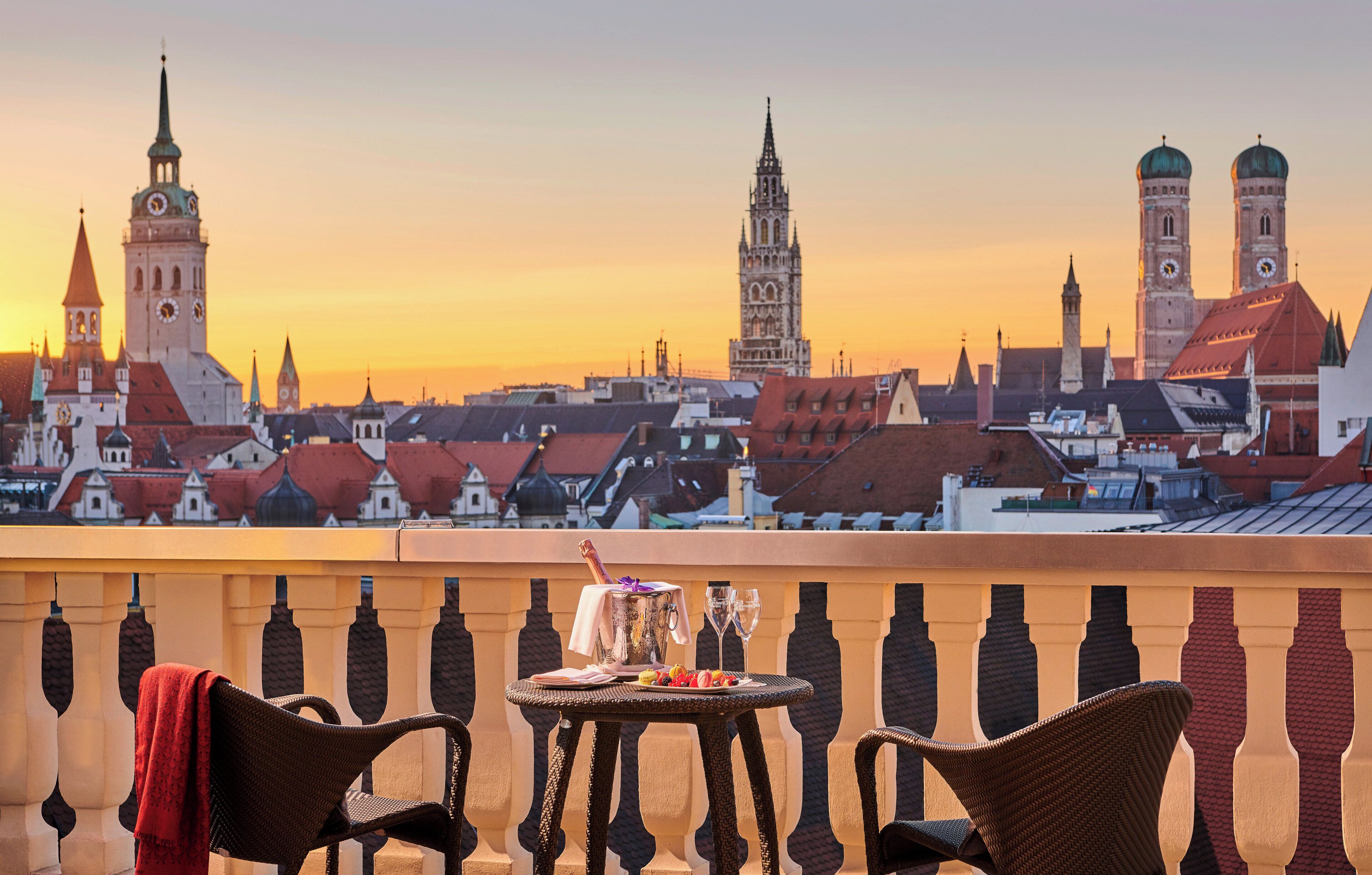 A balcony with wicker chairs, a table with a bottle and glasses, and a cityscape with historic buildings and a clock tower in the background.