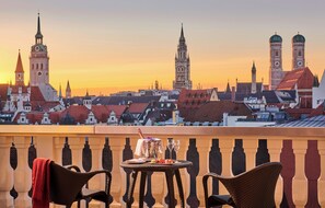 A balcony with wicker chairs, a table with a bottle and glasses, and a cityscape with historic buildings and a clock tower in the background.