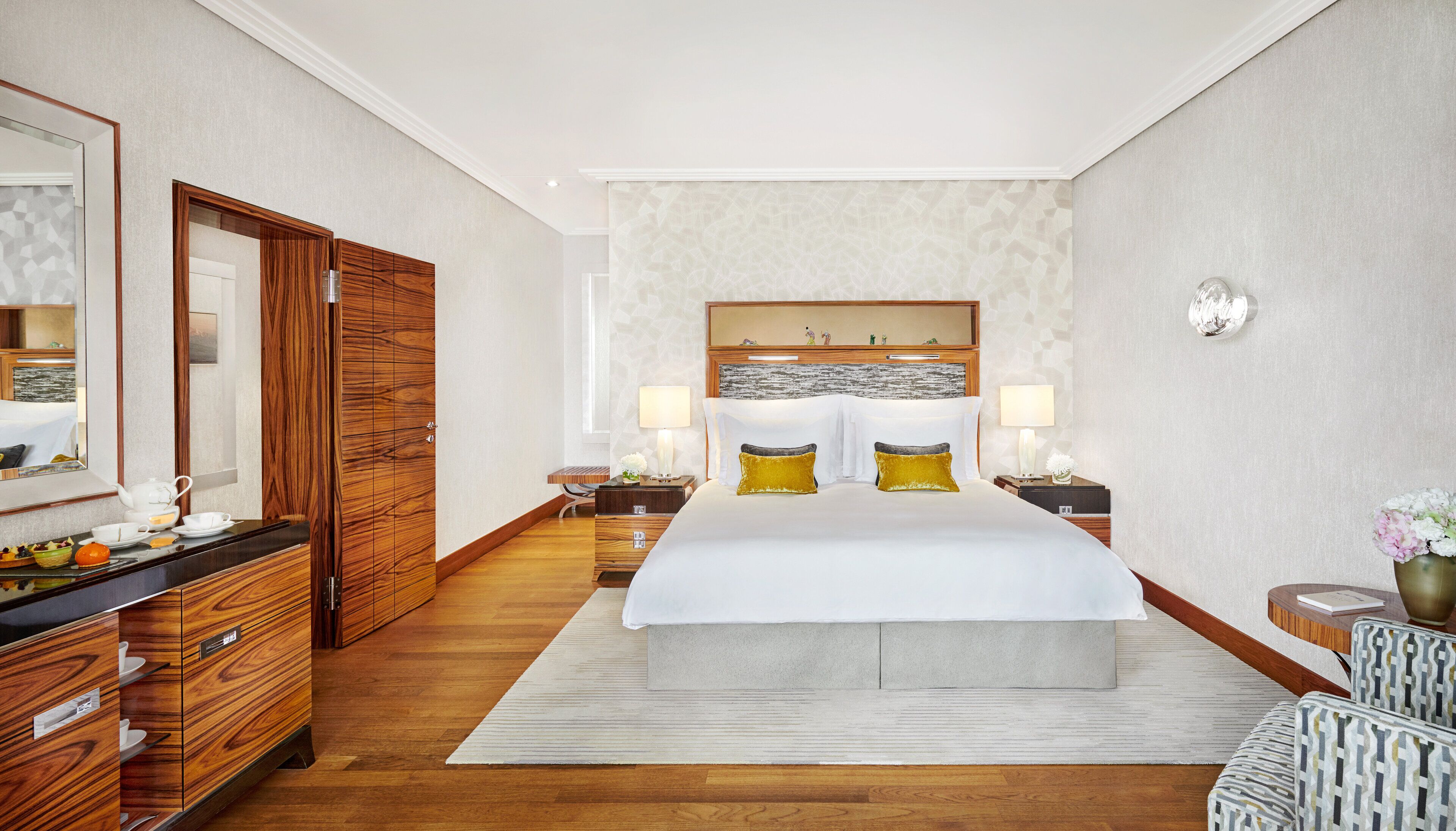 A hotel room with a large bed, wooden wardrobe, two bedside tables with lamps, a mirror, and a decorative headboard.