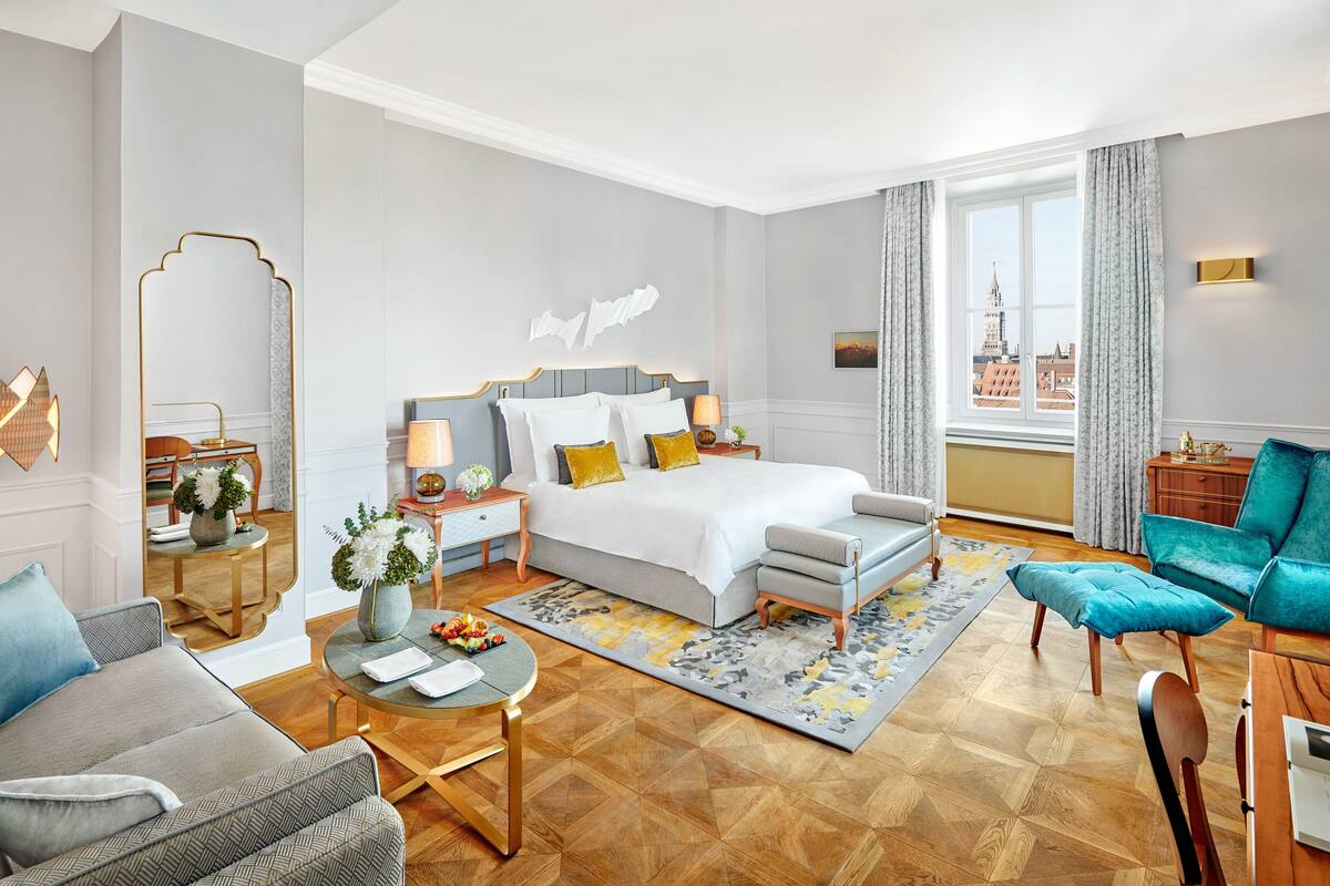 A modern hotel room with a large bed, a sitting area with a sofa and armchair, a dining table, and a view of a cityscape through the window.