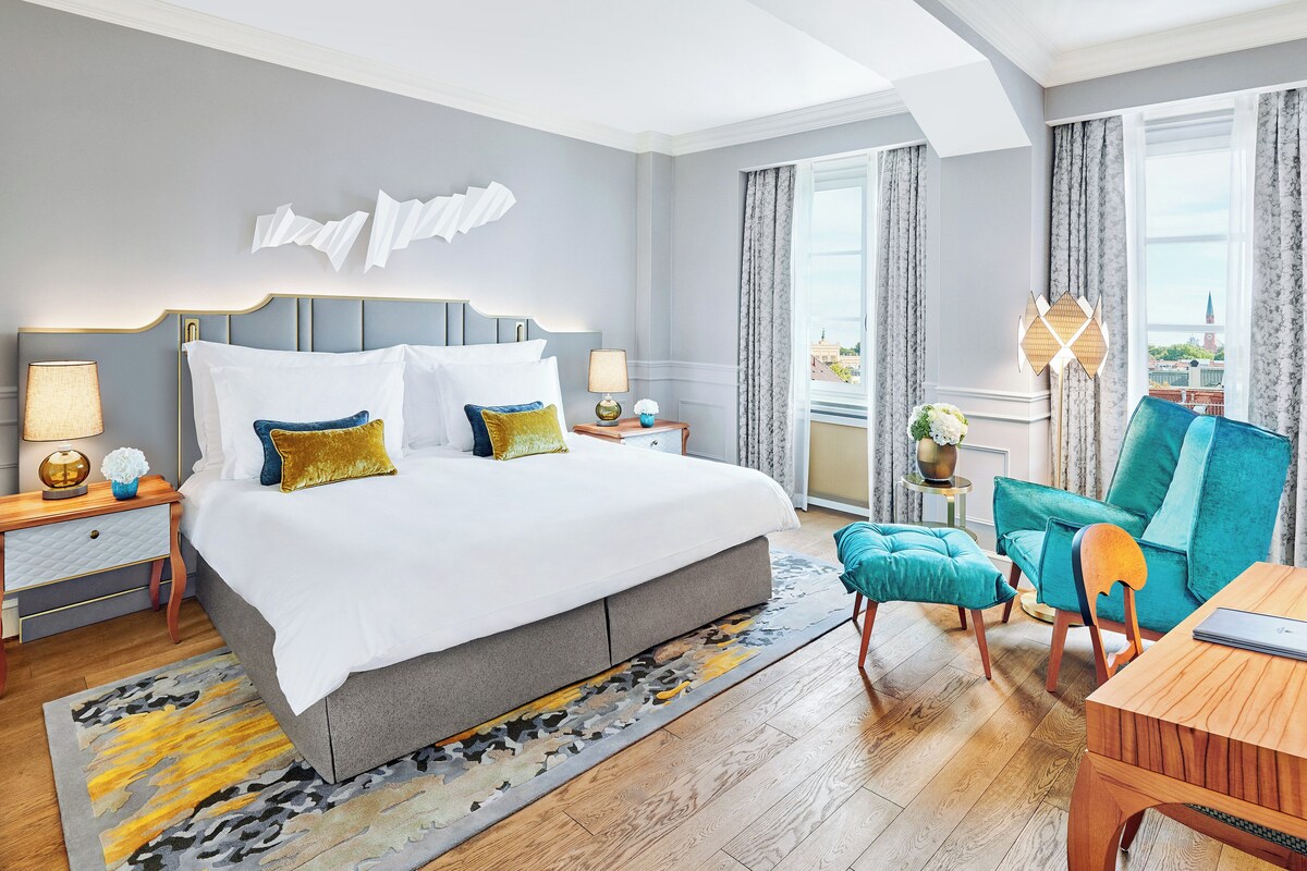 A modern hotel room with a large bed, a wooden desk, a teal armchair, and a view of the city.