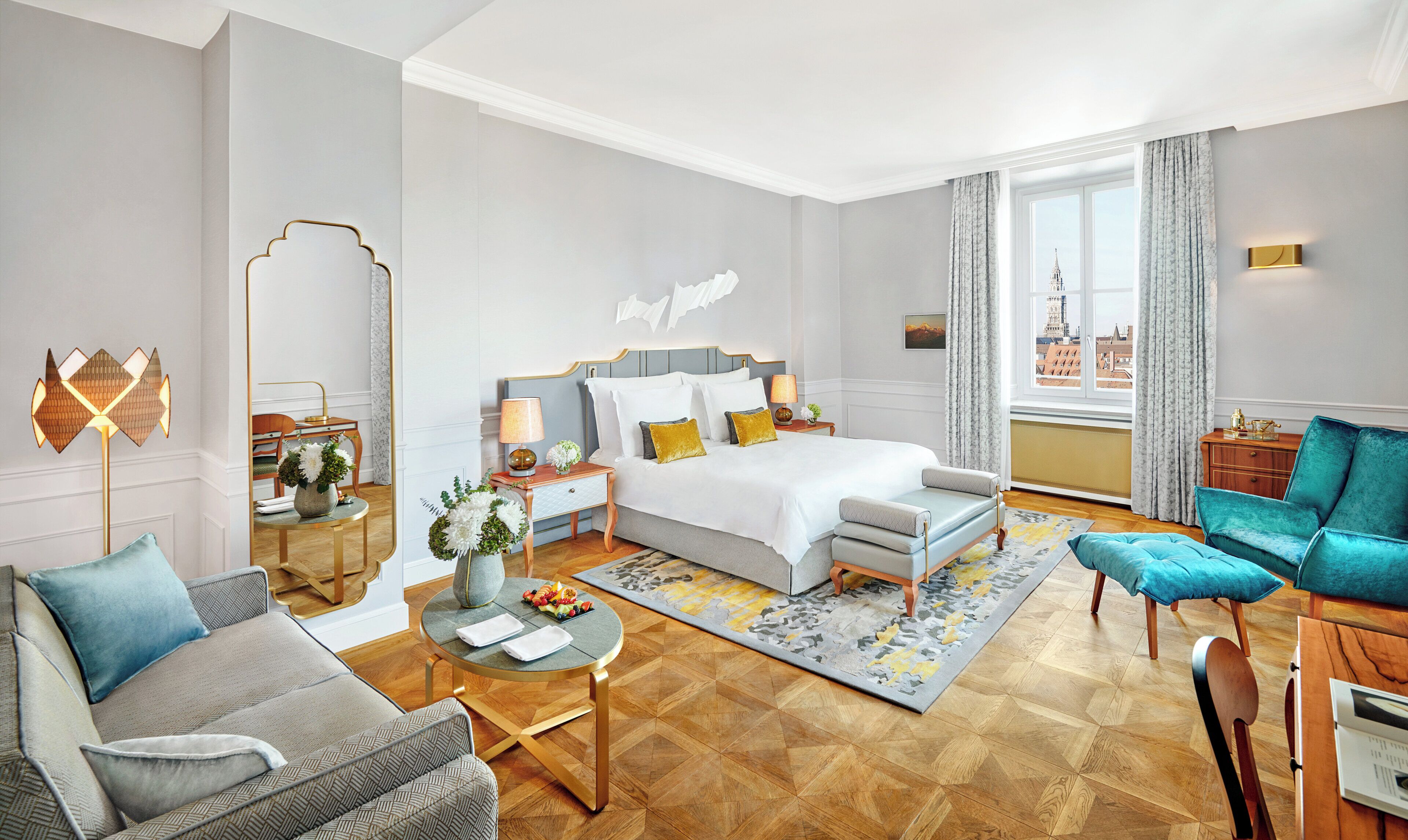 A hotel room with a bed, a seating area with a sofa and armchair, a dining table, and a view of a cityscape through the window.