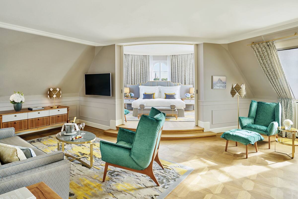 A spacious living area with a large bed, two green armchairs, a sofa, and a coffee table. A television is mounted on the wall, and there is a window with curtains.