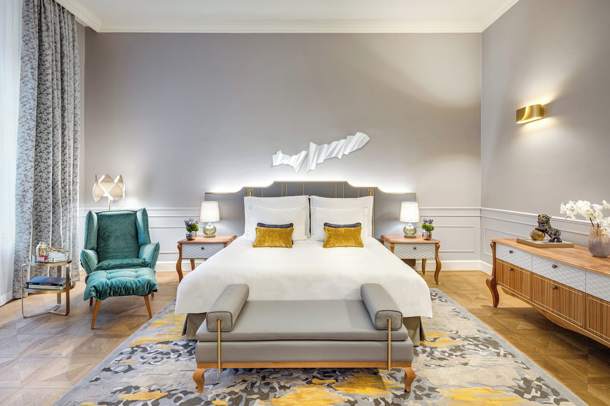 A modern hotel room with a large bed, a teal armchair, and a wooden nightstand with a lamp.