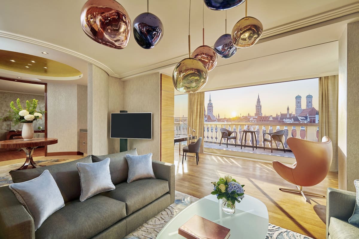 A modern hotel room with a sofa, a coffee table, and a TV. There is a balcony with a view of a city skyline.