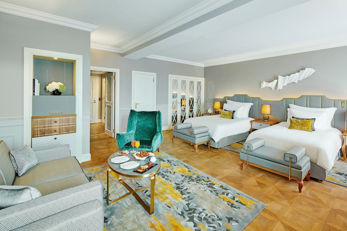 A hotel room with a large bed, two smaller beds, a sofa, a chair, a dining table with breakfast, and a decorative wall piece.