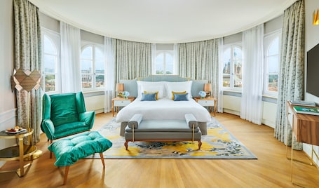 A spacious bedroom with a large bed, a green armchair, a side table, and a view of the city through large windows.
