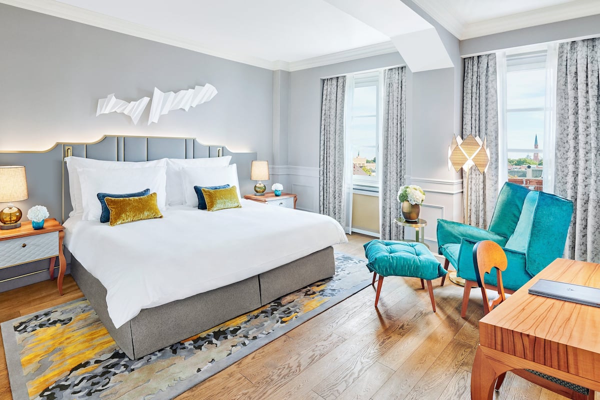A modern hotel room with a large bed, a wooden desk, a teal armchair, and a view of the city through large windows.