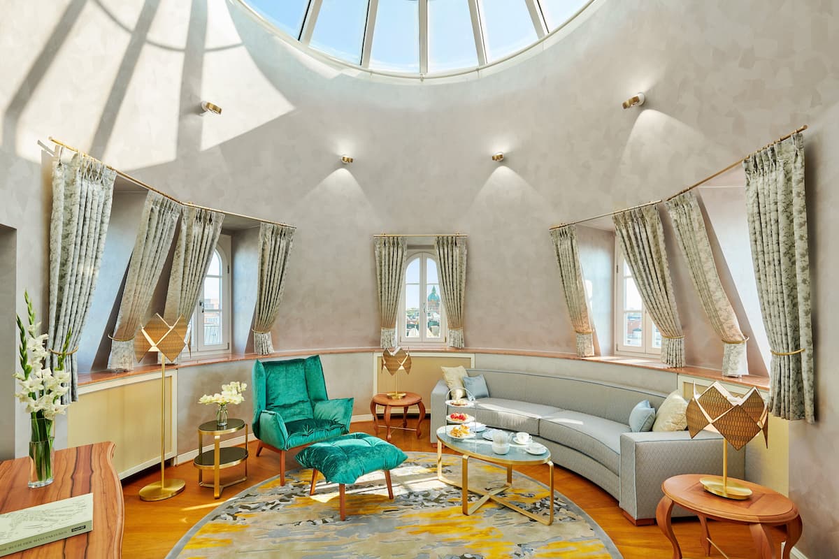 A spacious room with a curved sofa, a green armchair, a round coffee table, and a small side table. There is a large window with curtains and a skylight above.