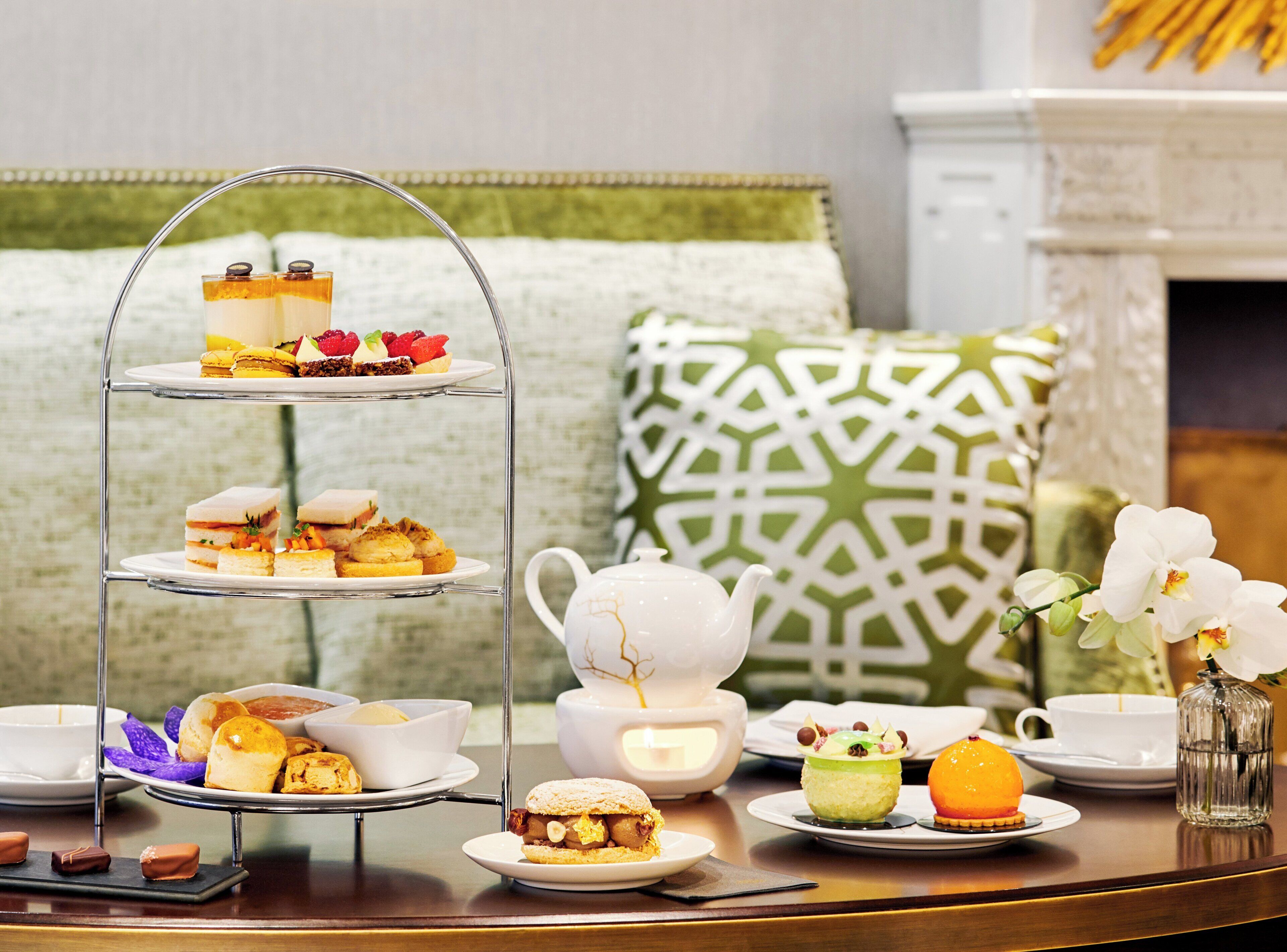 A three-tiered dessert stand with various pastries and a teapot on a table, surrounded by white cups and saucers.
