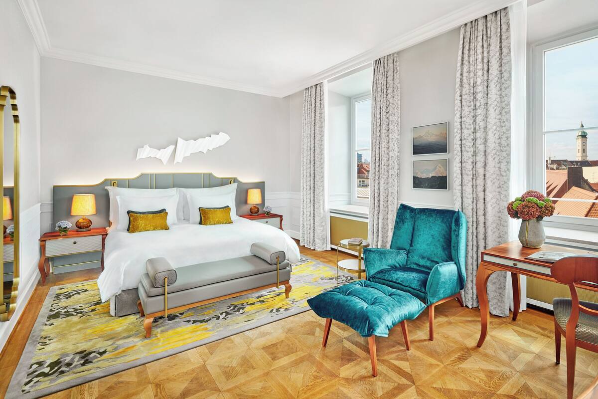 A hotel room with a bed, a blue armchair, a wooden desk, and a view of a cityscape.
