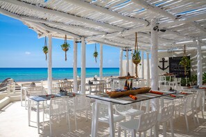 Un espace extérieur aménagé pour les repas, avec des meubles blancs, des plantes suspendues et une vue sur l’océan.