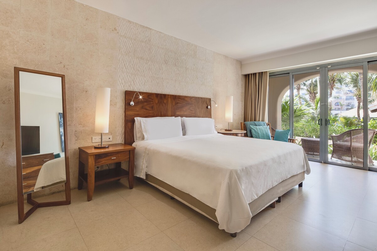 A hotel room with a large bed, a wooden nightstand, a mirror, and a view of the outdoors through a glass door.