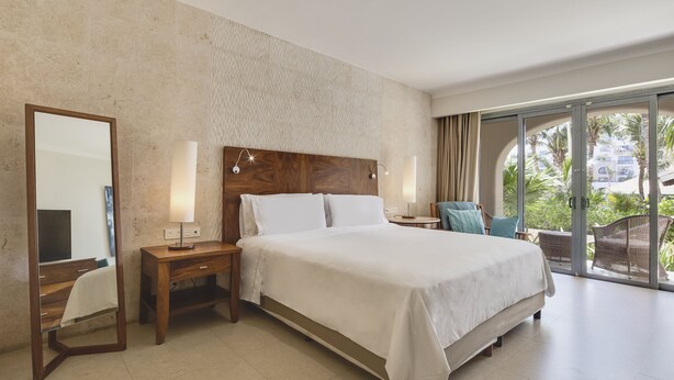 Ein Hotelzimmer mit einem großen Bett, einem Nachttisch aus Holz, einem Spiegel und Blick ins Freie durch eine Glastür.