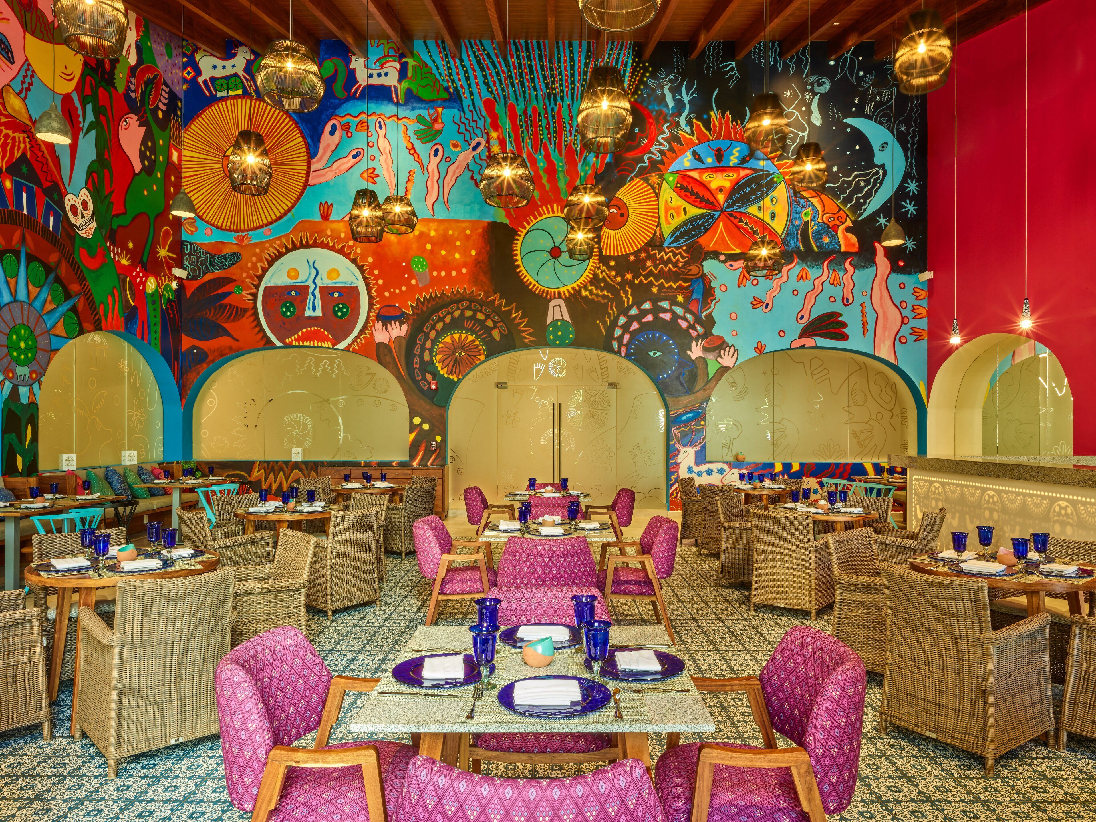 A vibrant restaurant interior with colorful murals, wicker furniture, and traditional table settings.